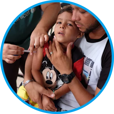 Foto mostra um menino recebendo uma vacina. Ele está sentado no colo do pai.