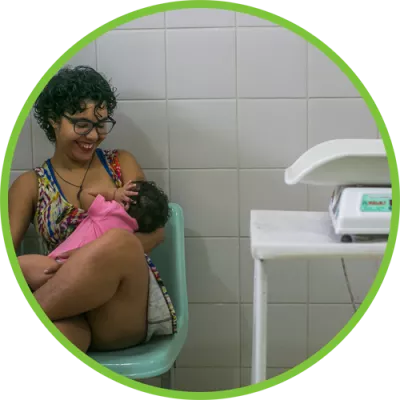 Foto mostra uma mulher sentada em um ambulatório médico ao lado de uma balança de bebês. Ela está segurando uma bebê no colo.