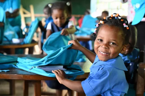 A girl smiles from her desk in a classroom while opening a UNICEF school kit