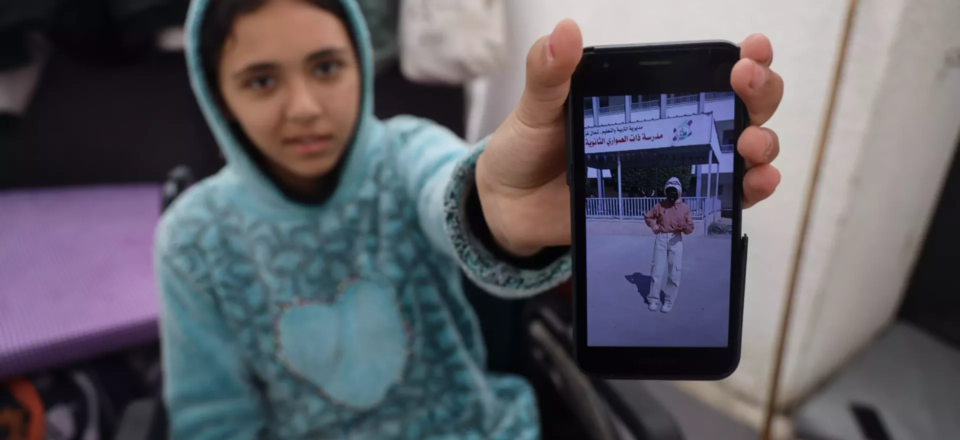Une jeune fille assise dans un fauteuil roulant tient son téléphone portable pour montrer une photo d’elle avant sa blessure.