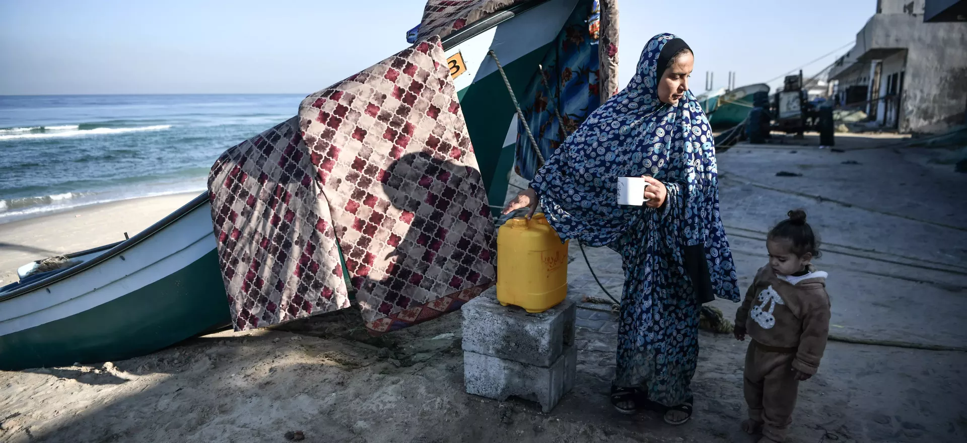 一位母亲和她蹒跚学步的女儿在海滩上的一艘渔船旁休息喝水。