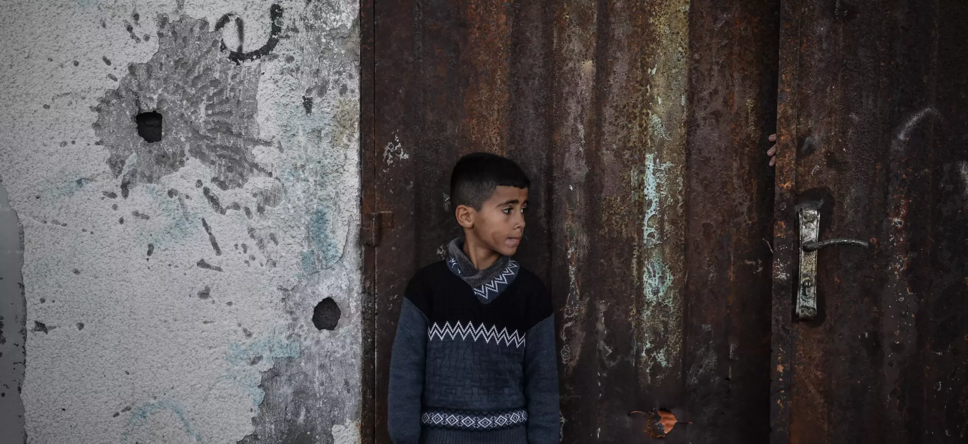 一名男孩站在一堵布满子弹的墙和一扇生锈的门旁边。