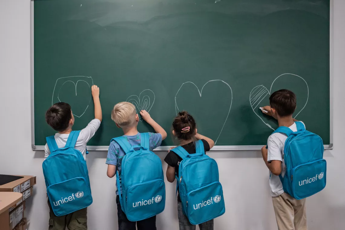 Des enfants de dos portant des cartables UNICEF dessinent des cœurs à la craie sur un tableau vert