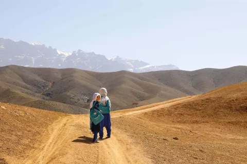  With mountains in the background, two girls walking on a dusty road look back. 