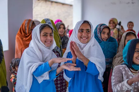 Two smiling adolescent girls in a class activity with their classmates in the background. 