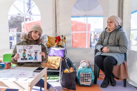 In a tent, a woman sits close to a girl showing a drawing to the camera. 
