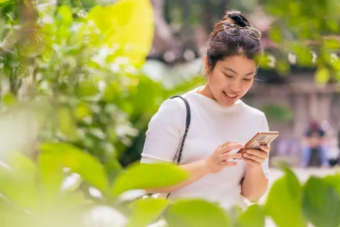 A smiling young woman with her hair tied up and glasses on her head is using a smartphone outdoors. She's dressed in a white elbow-length sleeve shirt and carries a black shoulder bag. She is surrounded by beautiful greenery, which slightly blurs the foreground, creating a feeling of depth and drawing attention to her activity with the phone.