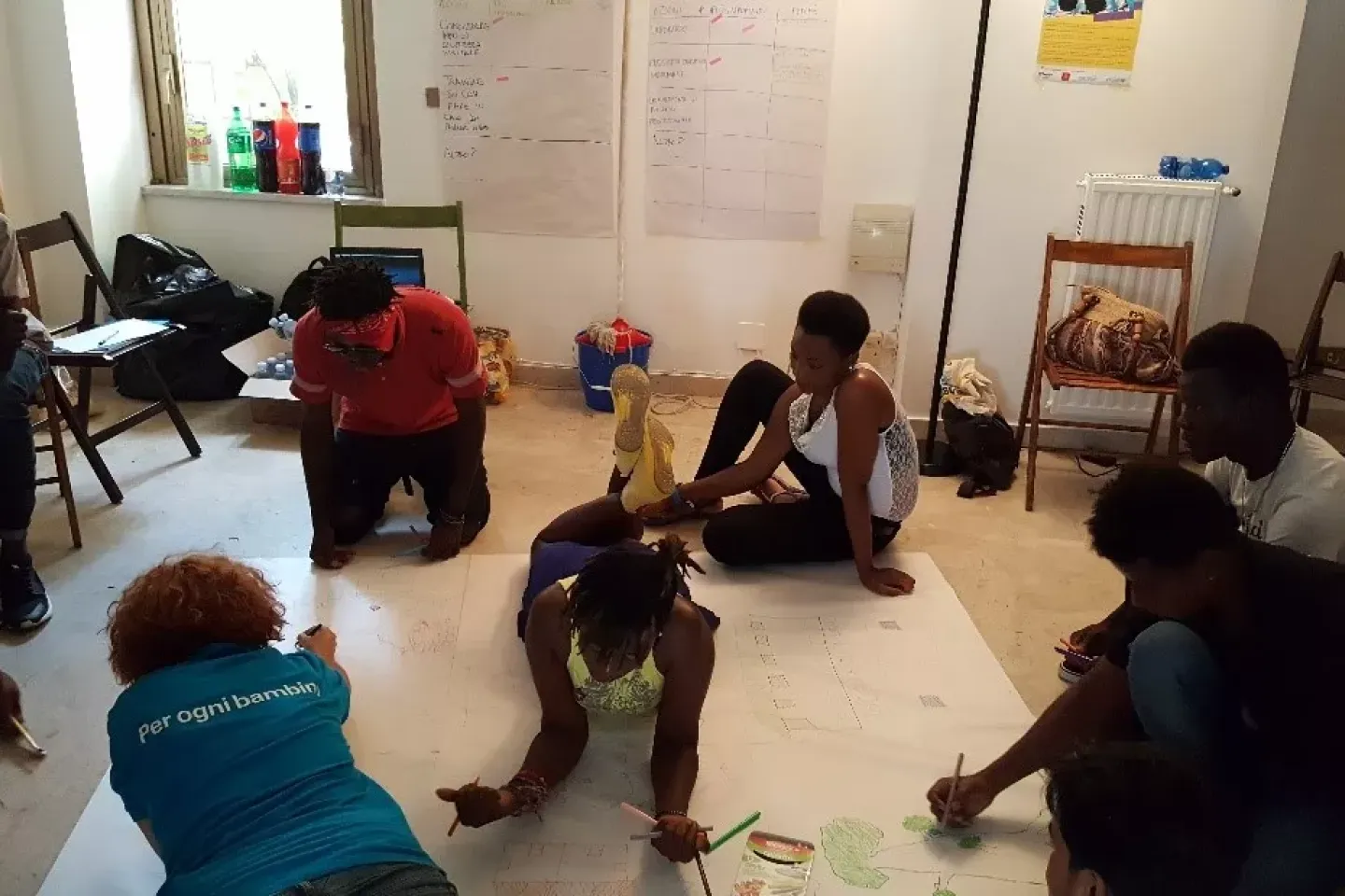 A group of young people sit around a large piece paper on the floor on which they draw and write.