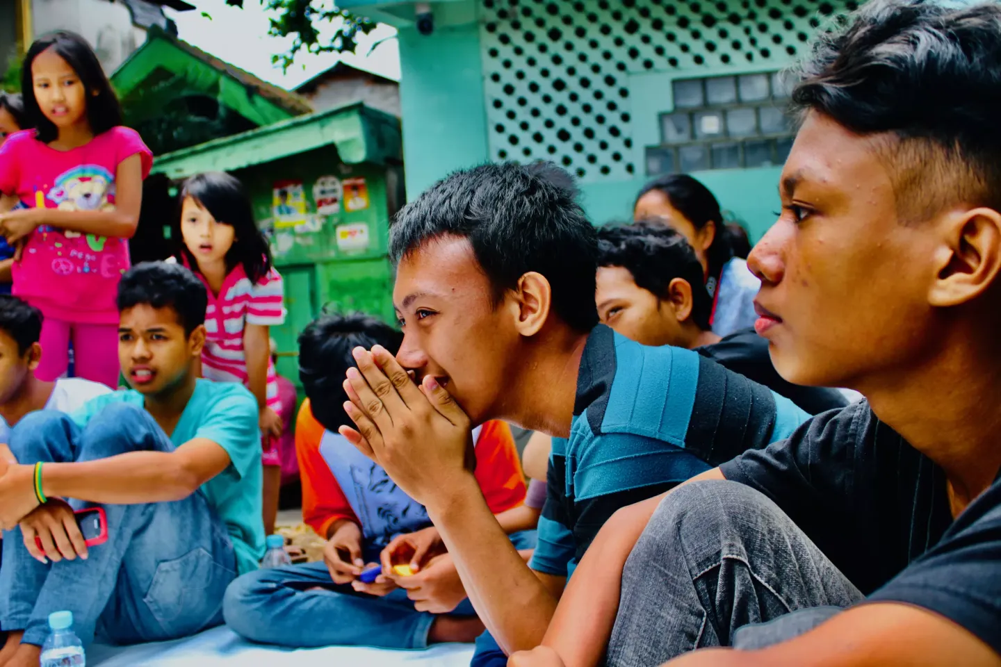 A group of adolescent girls and boys are standing up or sitting on the ground in an recreation activity in an urban area. Some are smiling and others are looking thoughtful.