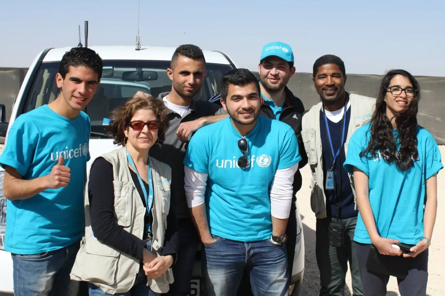 Four youth and three adults wearing UNICEF t-shirts, hats and vests are standing in front of a UNICEF jeep in a sunny outdoor place. They are looking at the camera and smiling.