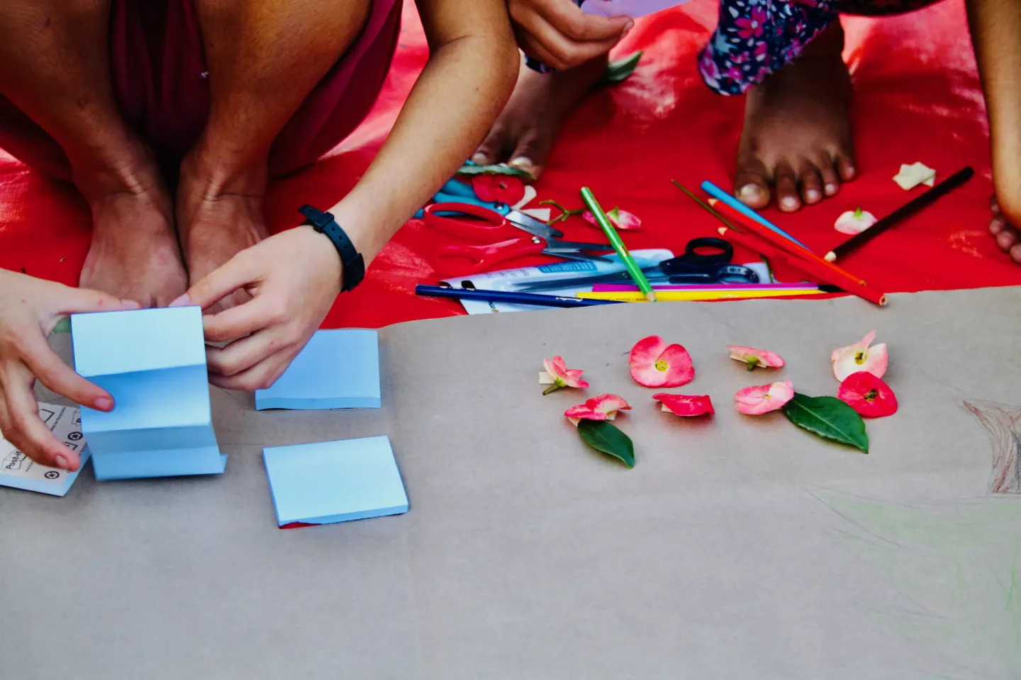 A piece of paper on the ground with flowers and smaller pieces of paper on it. Above it are two children's hands arranging items on it.