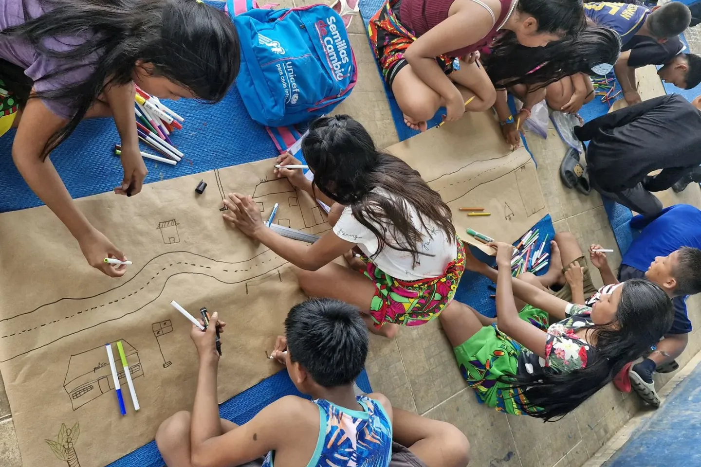 Several children are lying on their stomachs on a very large piece of brown paper and drawing. They are outside, and the photograph is taken from above them.