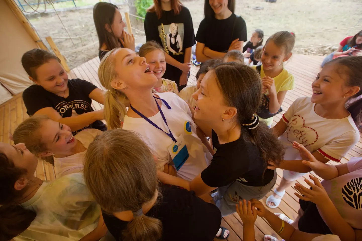 A group of girls and boys in Ukraine are standing around a young woman wearing a lanyard name tag. They are smiling and hugging her.