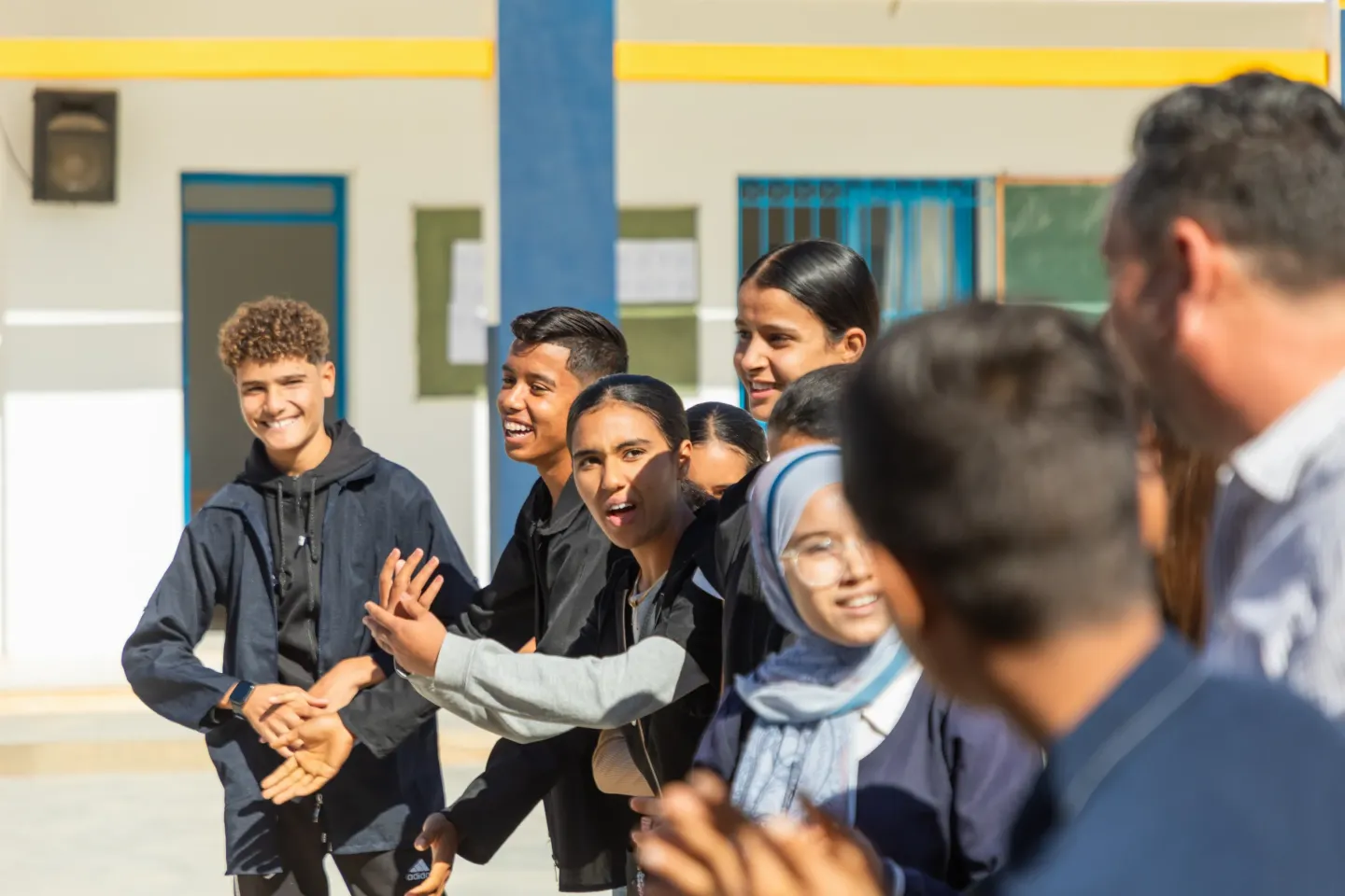 A group of adolescent boys and girls are standing in a line outside a school. They are laughing and smiling and facing in different directions