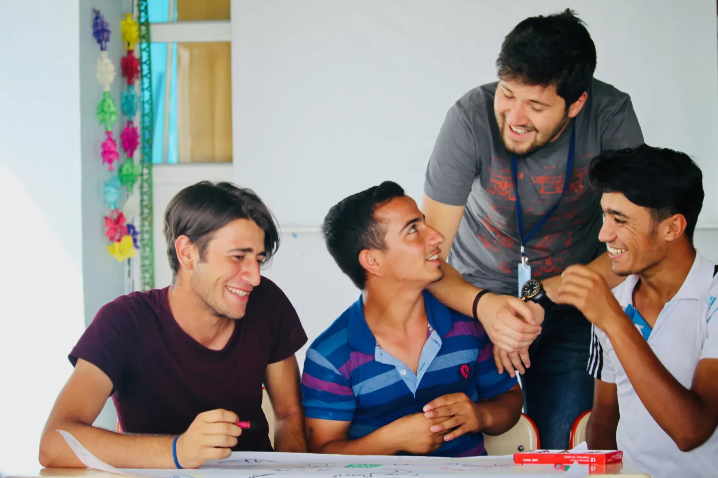 Three adolescent boys are sitting at a table with a piece of paper and pens resting on it. They are in a row facing the camera, and a young man is standing behind them, leaning over and talking with them. They are all smiling. 