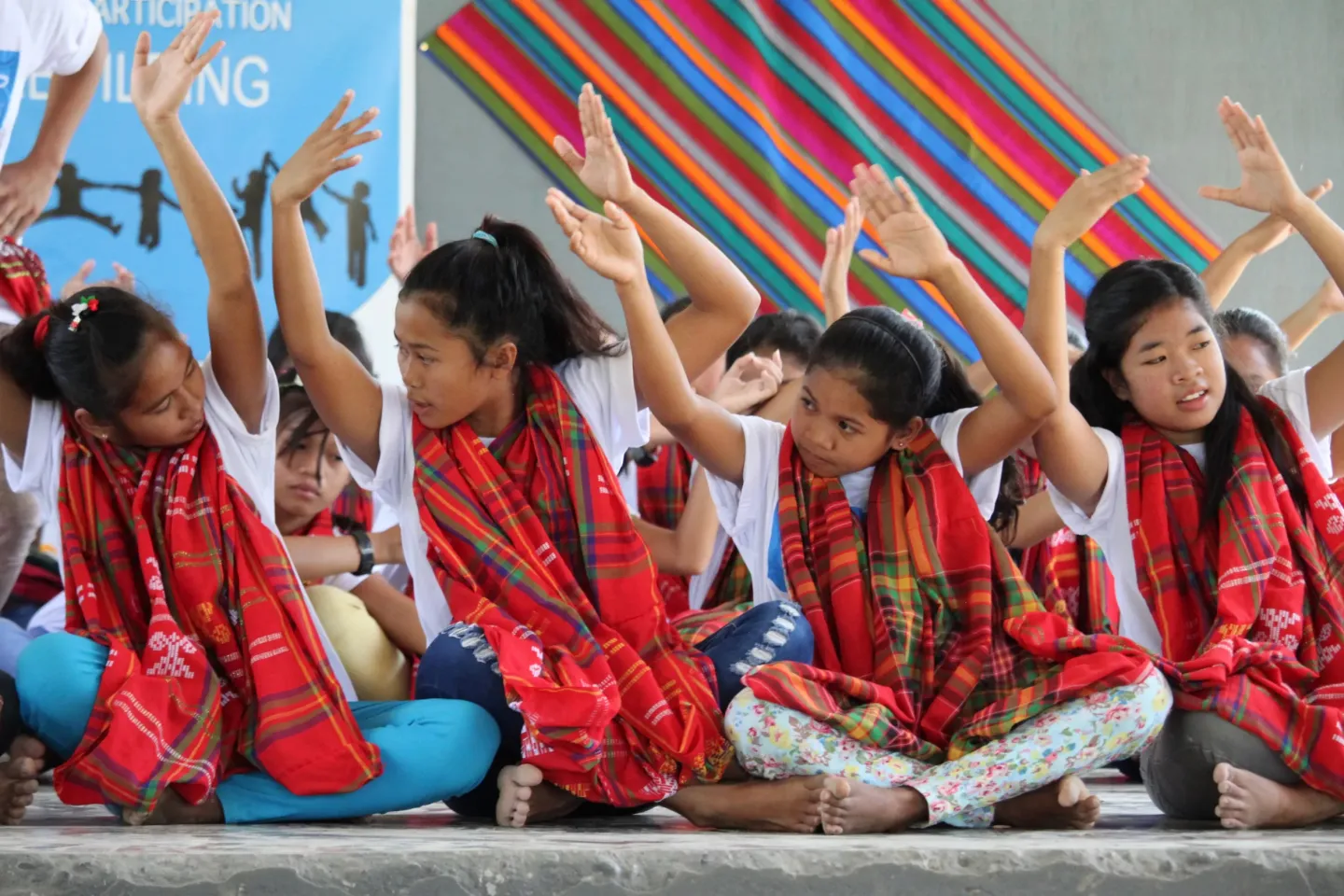 Teenage girls in the Philippines are sitting in a row crosslegged, holding their arms above their head in a traditional dance formation. They are wearing matching traditional outfits of red textile.