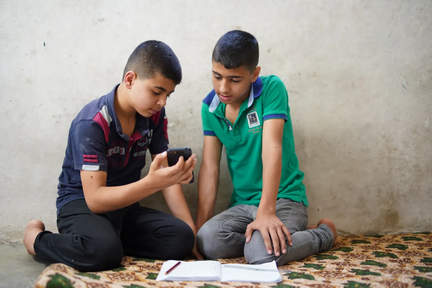 Two boys sit on the floor looking at a mobile phone with a book open on the floor in front of them.