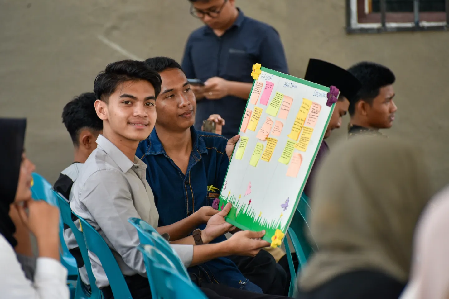 A youth smiles proudly while he holds a poster with colourful notes on it with his friend who is sitting next to him..