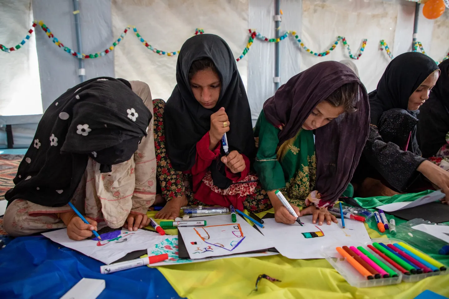 Young girls sit on the floor inside a tent drawing pictures.