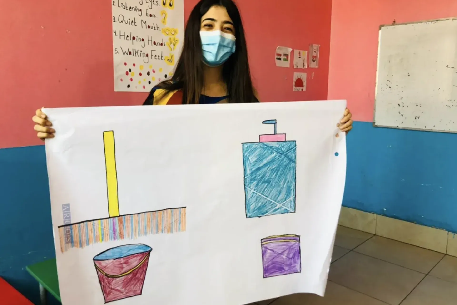 An adolescent girl wearing a surgical mask is holding a large piece of paper in front of her with colorful drawings on it. She is standing in a room with colorful drawings and paint on the walls.Her eyes appear to express a smile.