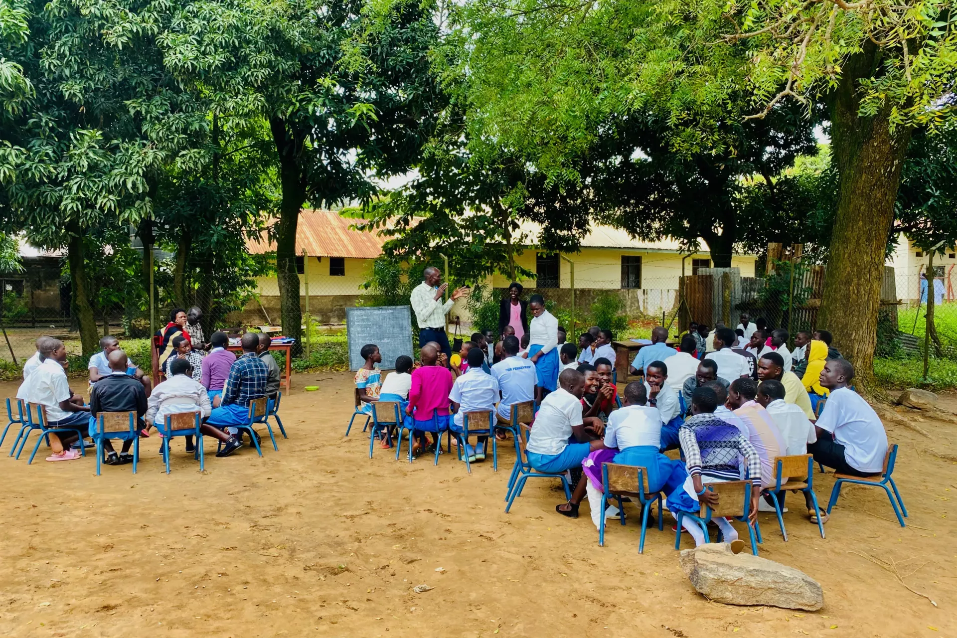 Secondary school students are sitting on chairs arranged in small circles in a schoolyard in Uganda. Two teachers, one male and one female, are standing and walking between the groups.