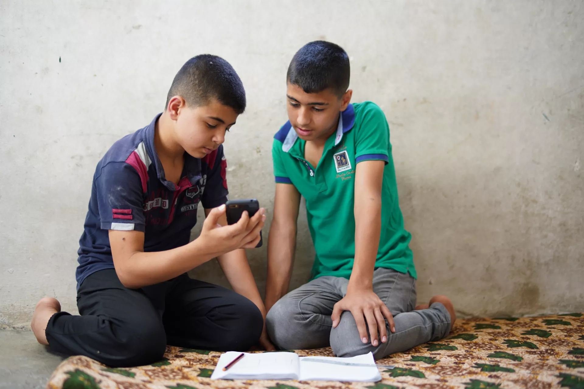 Two boys sit on the floor looking at a mobile phone with a book open on the floor in front of them.