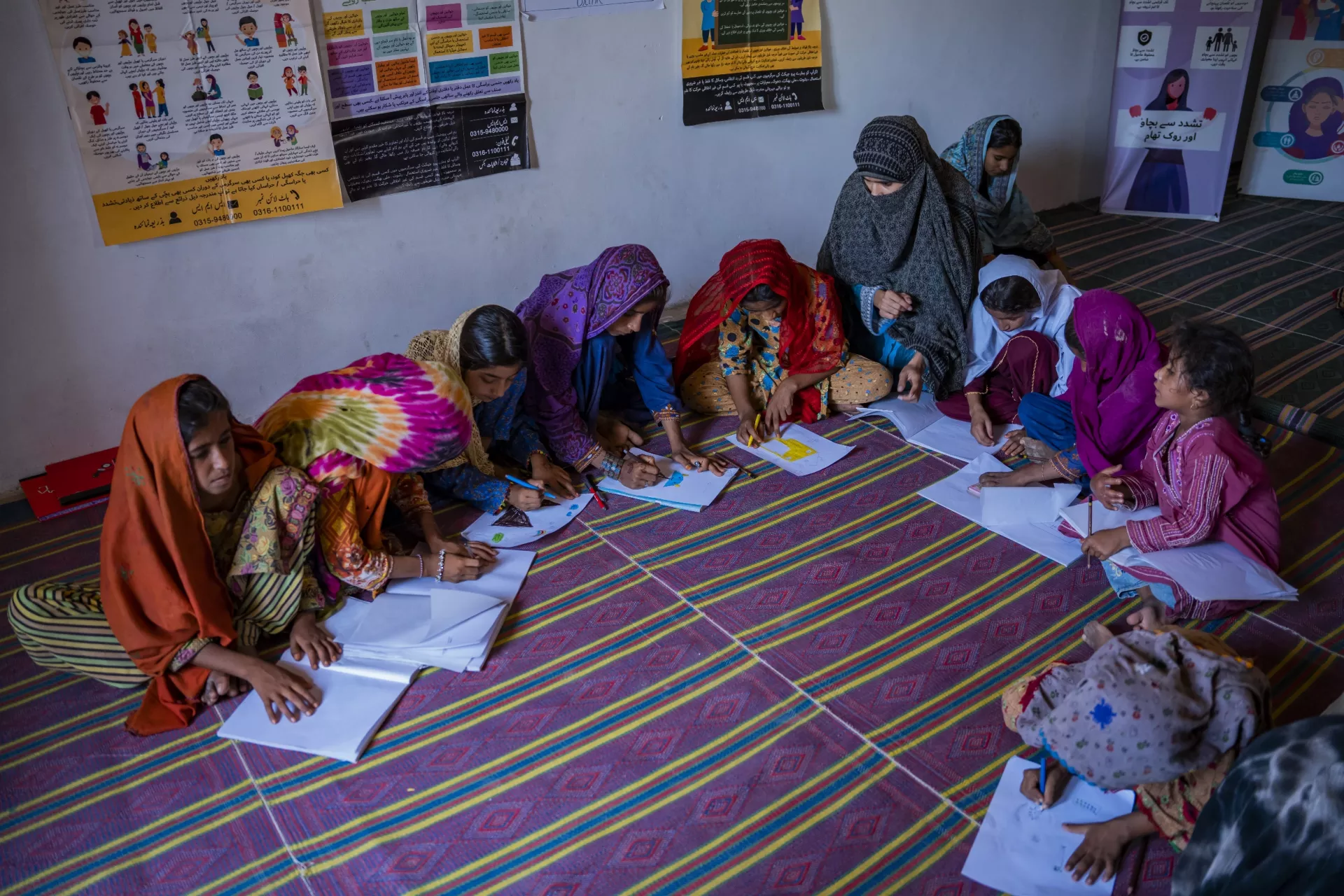 A group of children sit on a carpet inside making drawings on paper in front of them on the floor.