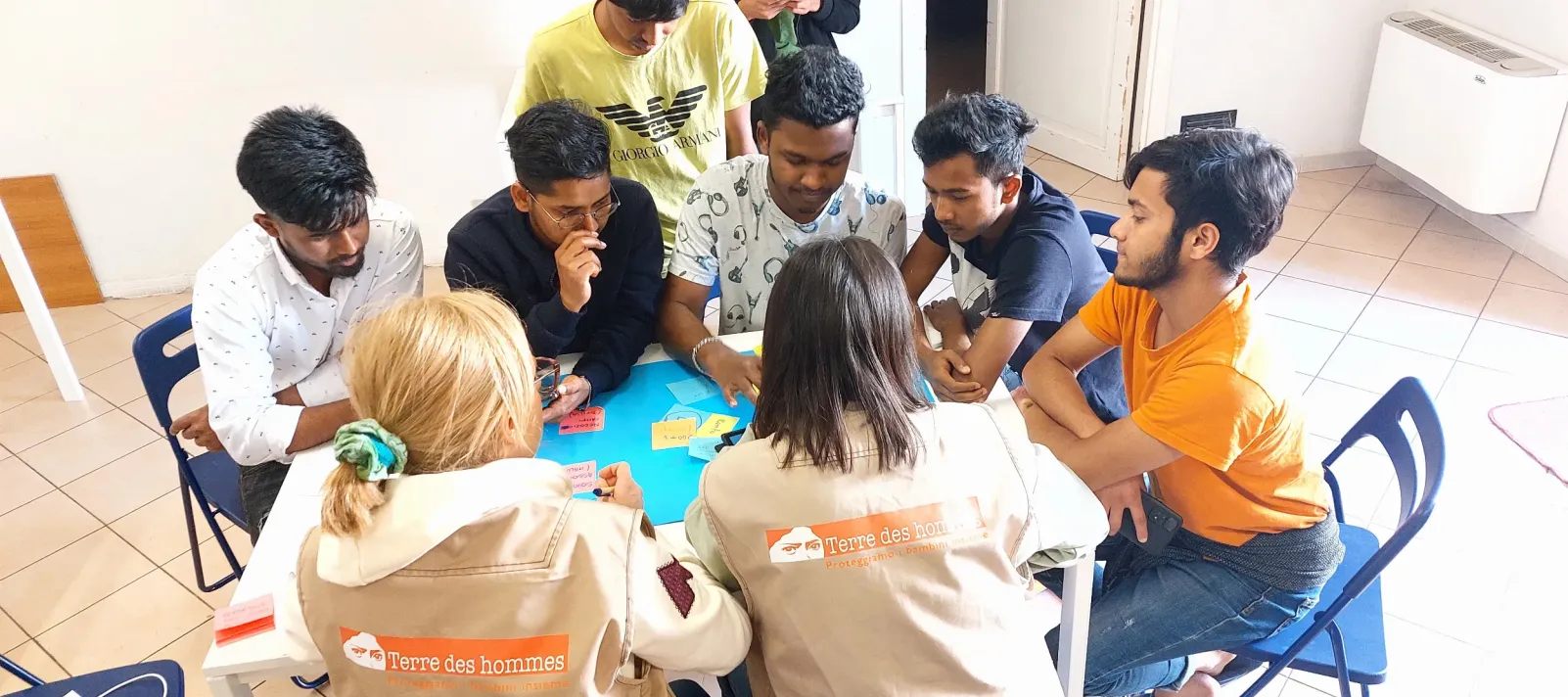 A group of adolescent boys are sitting around a table looking at colorful cards with handwriting on them. Two adullts wearing field vests with the name "Terre des hommes" are sitting at the table with their backs to the camera. They are in a classrom or activity space.