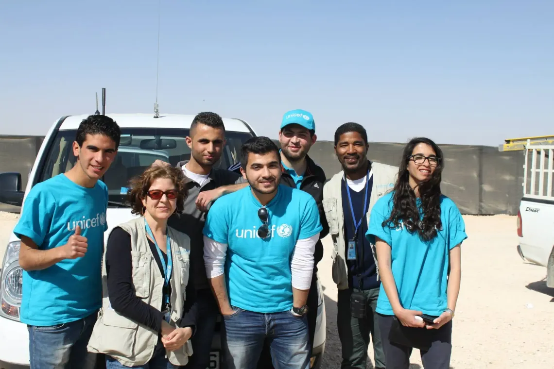 Four youth and three adults wearing UNICEF t-shirts and vests are standing in front of a white UNICEF jeep at the entrance of a refugee camp in Jordan. They are looking at the camera annd smiling.
