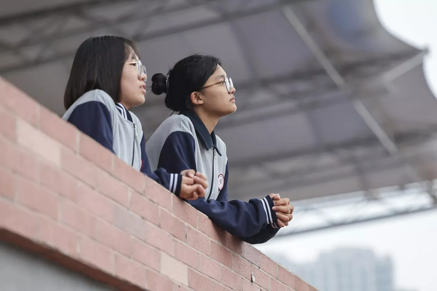 Two adolescent girls, in school uniforms, look out a brick wall of their school