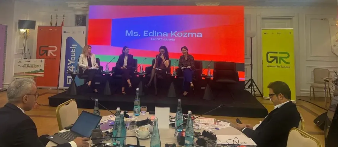 Four speakers on stage during a panel, colorful screen backdrop, water bottles on tables in the foreground.