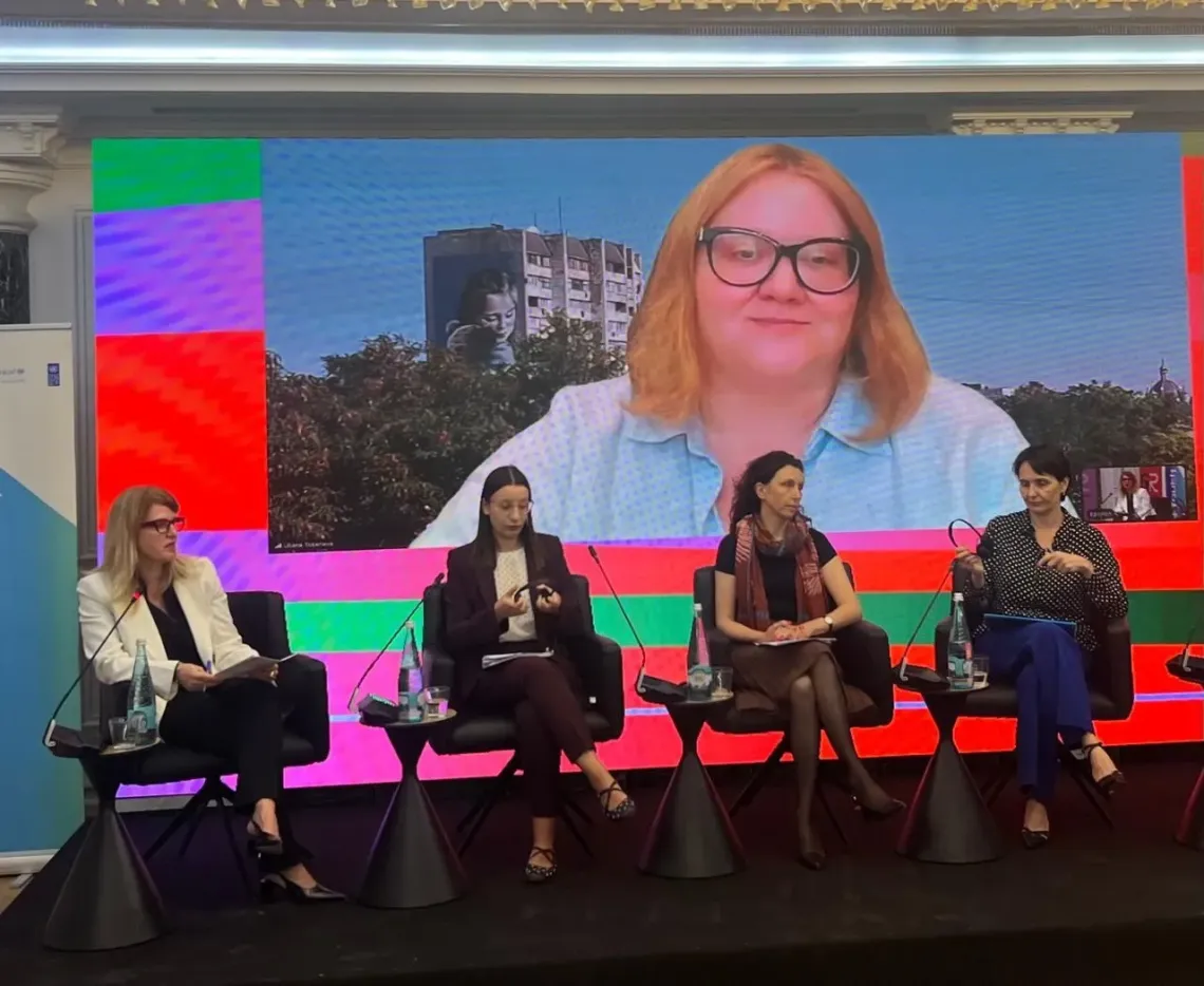 Four panelists seated on stage with microphones; large screen behind displays a remote speaker and colorful backdrop.