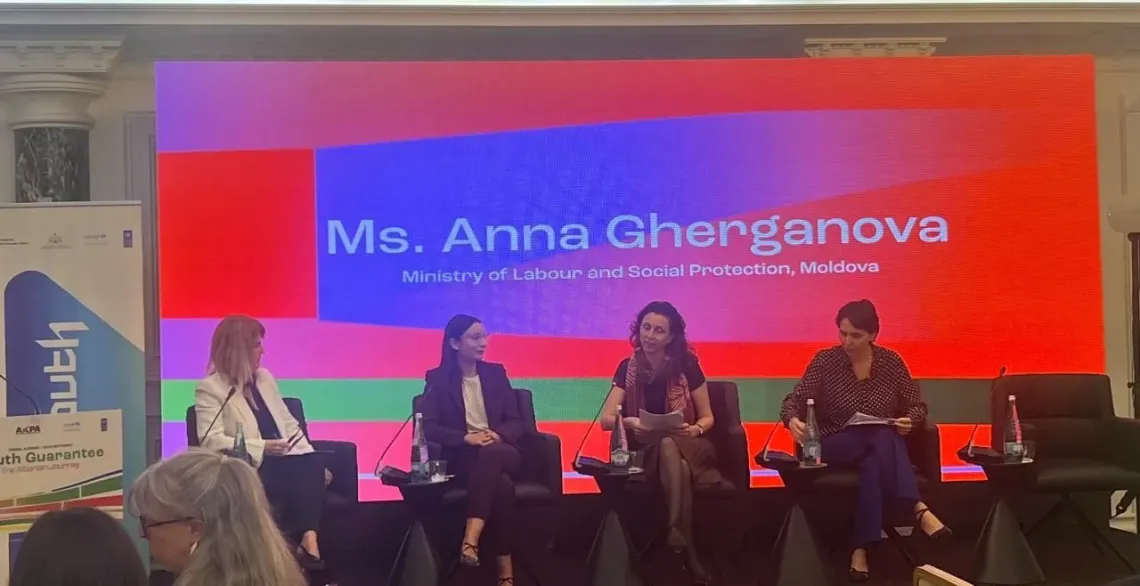 Panel discussion with four speakers on stage in front of a bright, multicolored presentation screen.