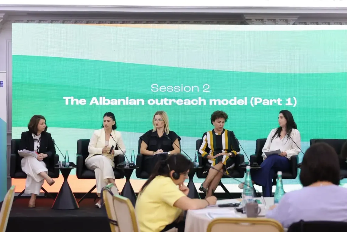 Panel of five speakers on stage discussing the Albanian outreach model while audience listens in a conference hall.