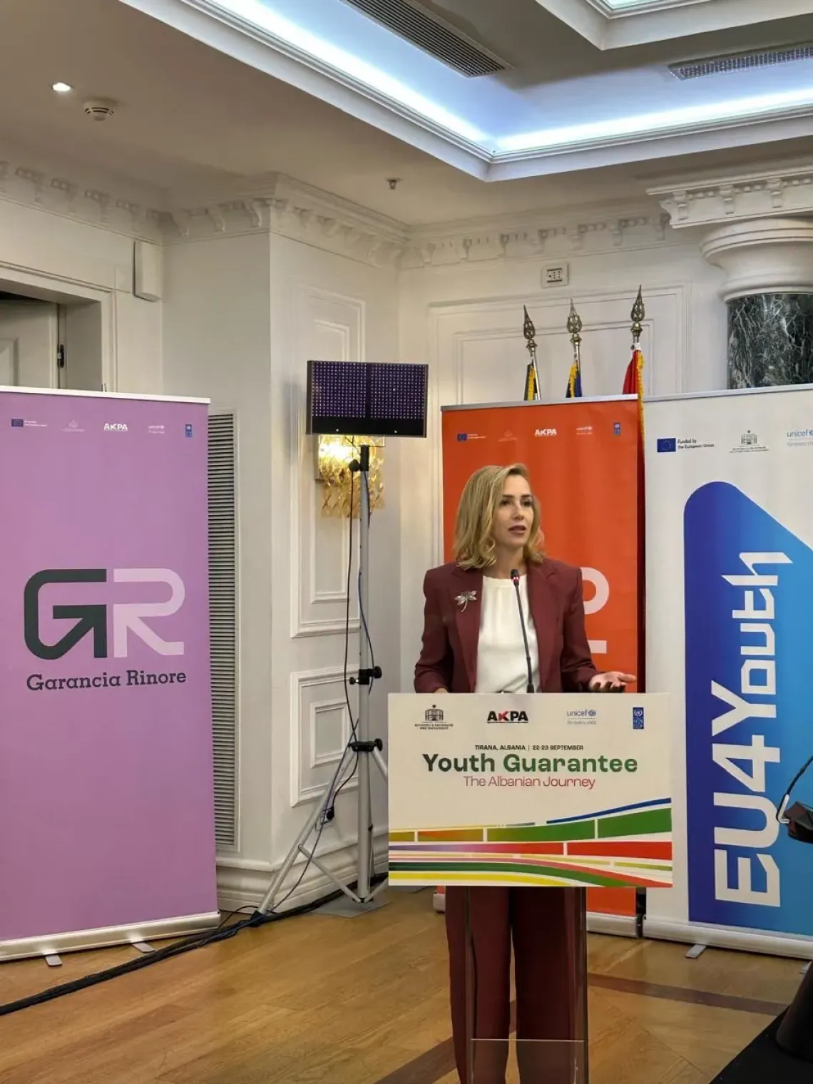 Deputy minister Olta Manjani at podium during EU4Youth event, flanked by banners and flags, presenting on Youth Governance.