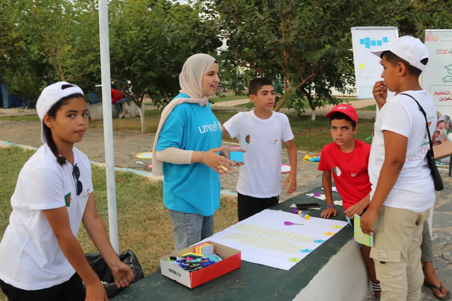 Children take part in an outdoor workshop around a table covered with papers, markers and sticky notes, while a facilitator leads the discussion.