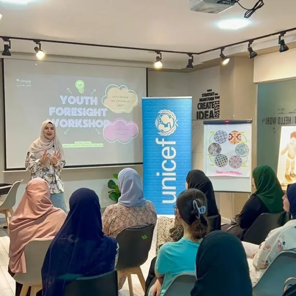A facilitator addresses seated participants during a Youth Foresight workshop, with a UNICEF banner, a presentation screen and workshop materials visible at the front of the room.