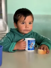Ni&ntilde;o con abrigo verde tomando desayuno con una taza y un pan en la mano