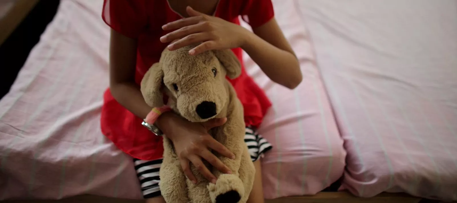 Una ni&ntilde;a sentada en el borde de una cama sostiene su perro de peluche y mira hacia abajo.
