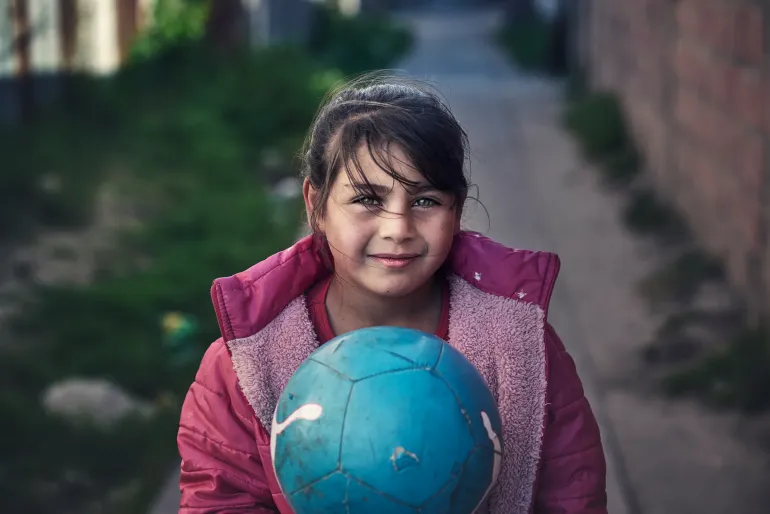 Una ni&ntilde;a en un barrio vulnerable mira a c&aacute;mara sosteniendo una pelota de f&uacute;tbol.