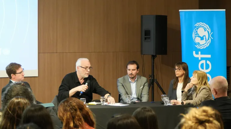 Mesa redonda con tres panelistas frente a p&uacute;blico; un hombre mayor habla al micr&oacute;fono.