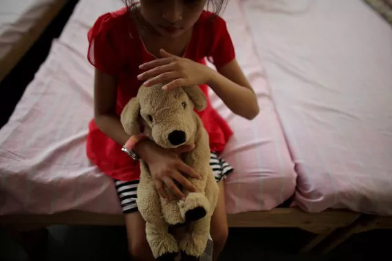Una ni&ntilde;a sentada en el borde de una cama sostiene su perro de peluche y mira hacia abajo.