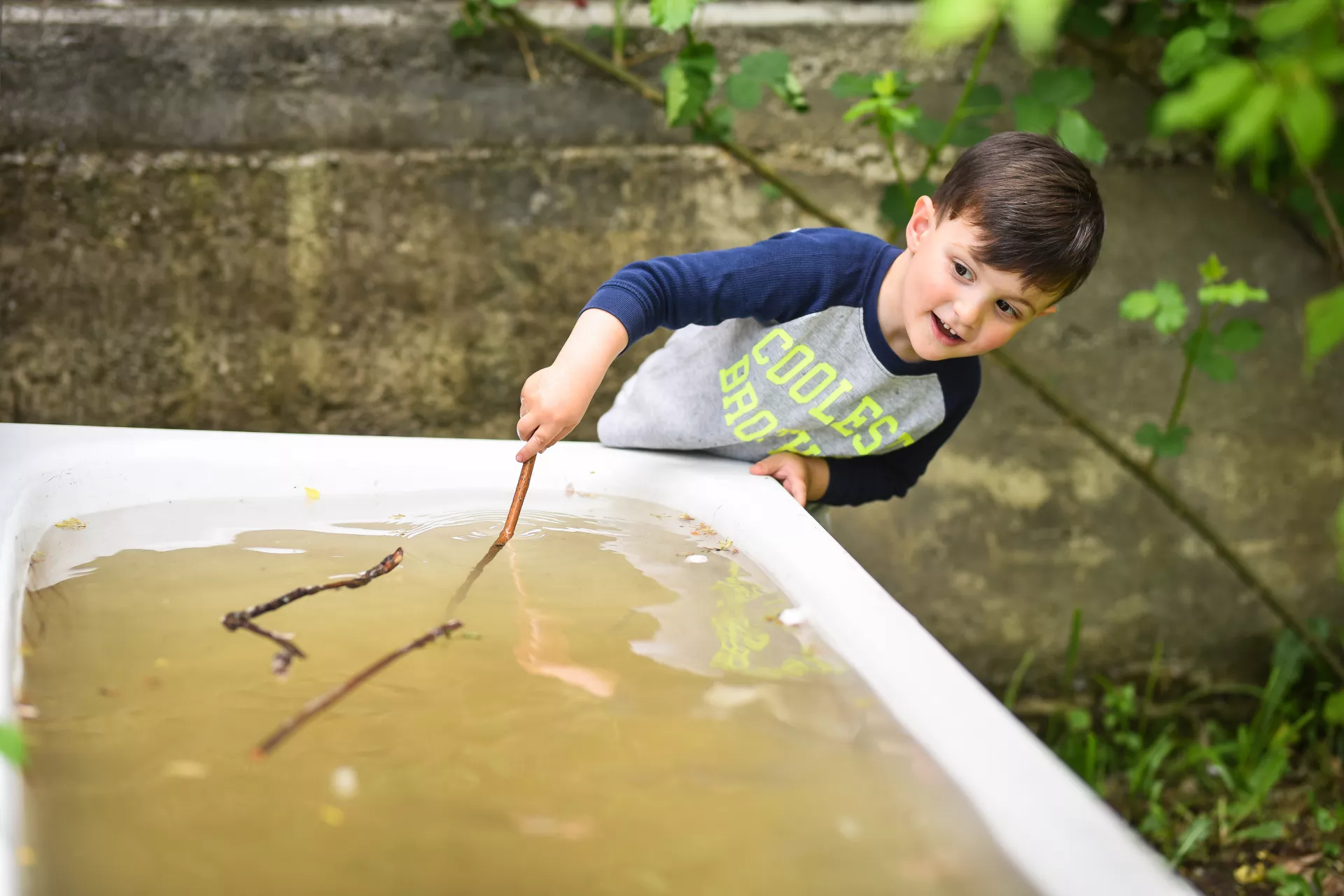 A boy with a stick is playing with collected water in a tab outside