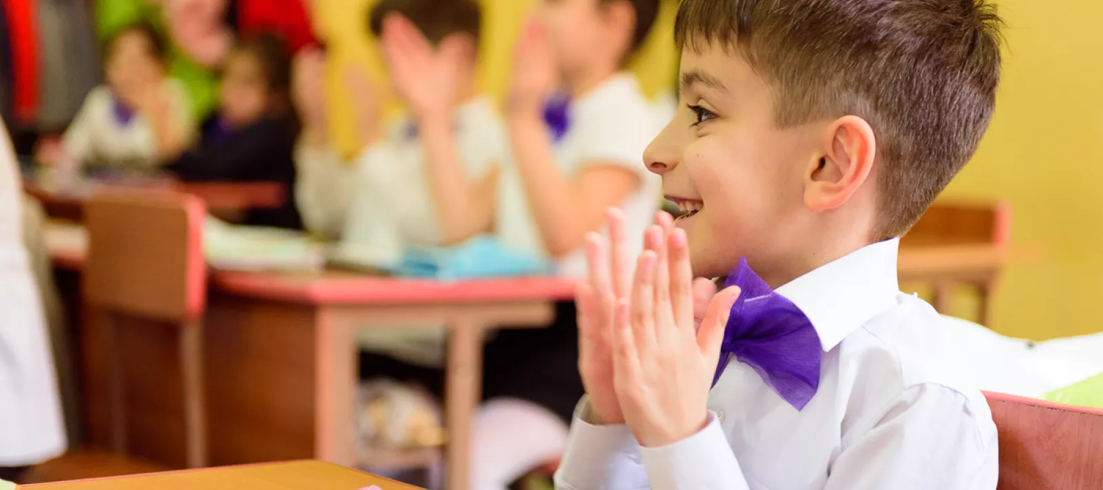 A boy with a purple bow over his white shirt claps his hands and smiles in the classroom during the lesson.
