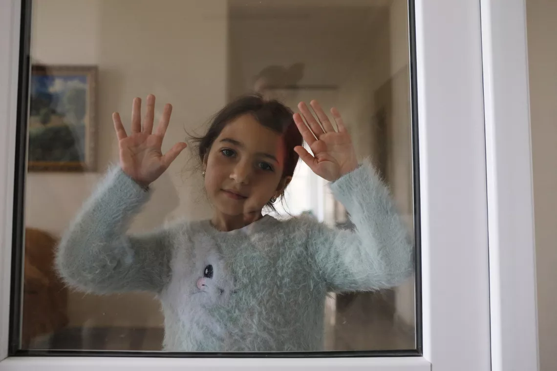 A young girl posing from the camera with two hands placed on the window.