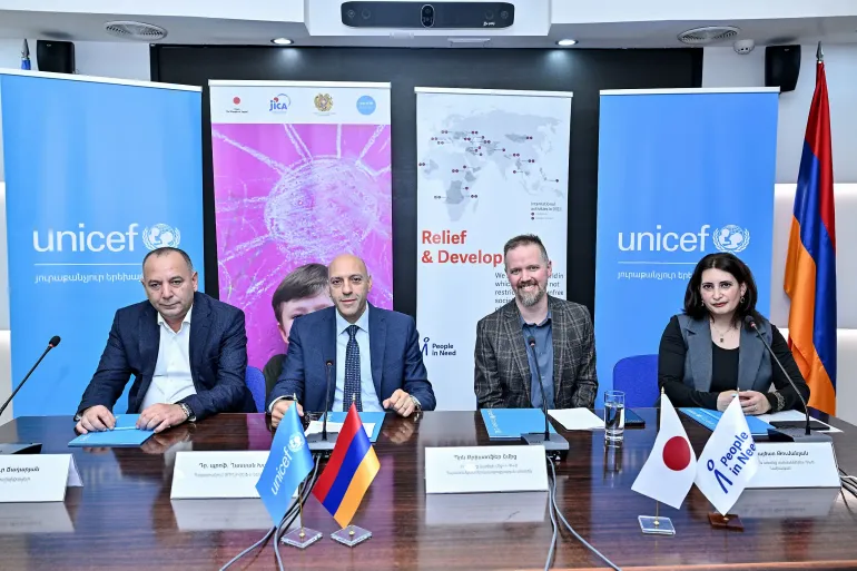 Four officials seated at signing table with UNICEF and partner banners, UN, Armenia, and Japan flags displayed.
