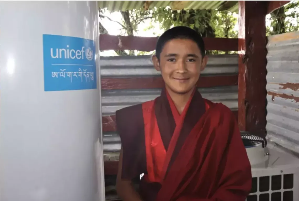 Young monk stands next to a water heating system with UNICEF written on it.