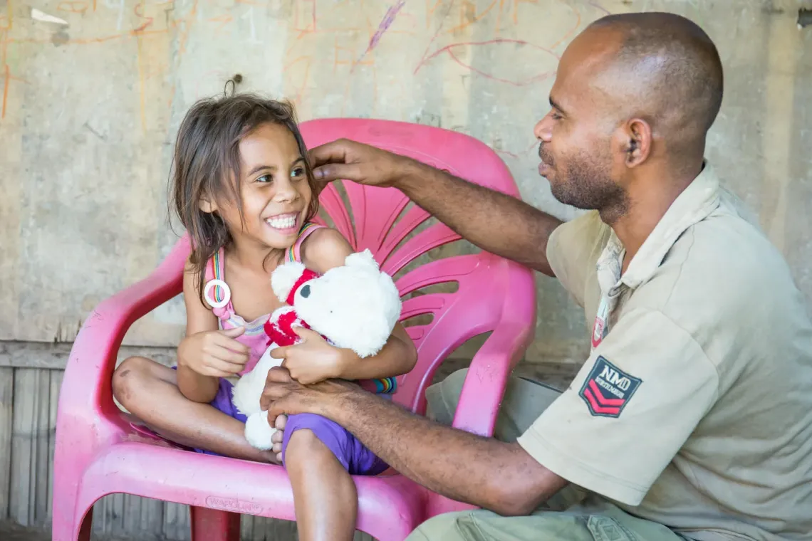 A girl sits in a pink chair holding a stuffed animal smiling at her father