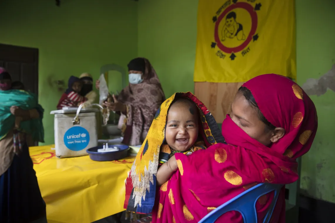 a child smiles over her mother's shoulder with health workers in the background