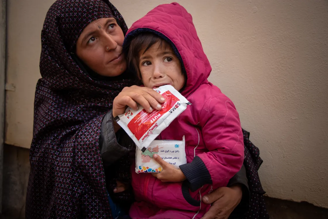 a toddler sits in her mother's lap while she eats from s ready-to-use therapeutic food packet